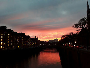 Silhouette buildings by river against sky at sunset