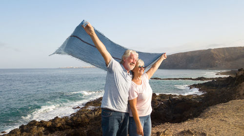 Happy senior couple holding textile while standing on rocky coastline