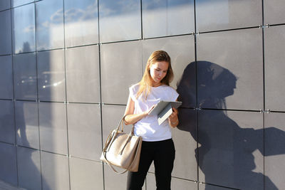 Young businesswoman using digital tablet standing against wall