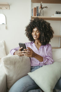 Smiling woman using smart phone sitting on sofa at home