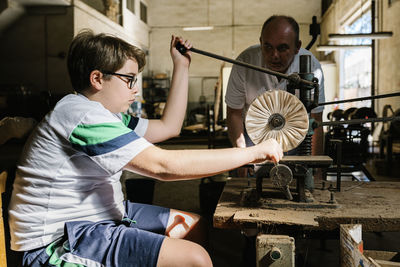 Focused teenage boy working with old drill press under supervision of father while polishing detail in workshop