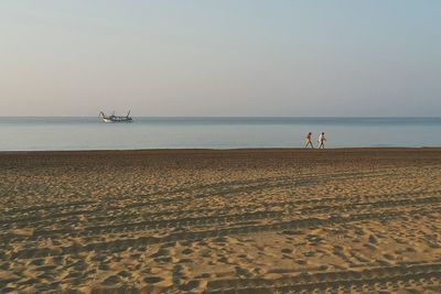 Distant view of friends walking at shore against clear sky during sunset