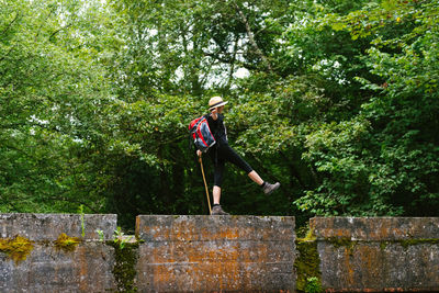 Side view of unrecognizable relaxed female tourist with backpack crossing calm river while walking along stone fence with outstretched arms