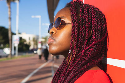 Side view portrait of young african american woman in braids and trendy outfit with sunglasses looking away while standing against red background in street