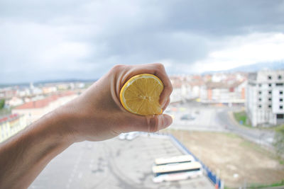 Cropped hand of man squeezing lemon against cityscape