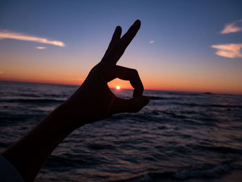 Silhouette hand gesturing ok sign over sea against sky during sunset