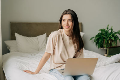 Attractive thoughtful female student sitting on bed and using laptop for communication