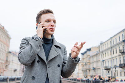 Emotional young man using mobile phone, wearing an elegant gray coat, stands