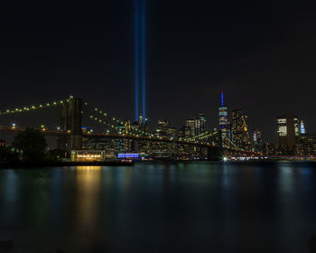 Illuminated bridge over river by buildings against sky at night