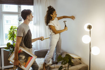 Woman hammering nail in wall with boyfriend holding picture frame and measuring tape at home