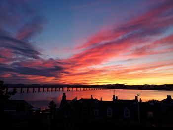 Silhouette buildings by sea against dramatic sky during sunset