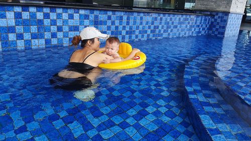 Mother teaching her child to swim with a yellow inflatable ring in a blue swimming pool