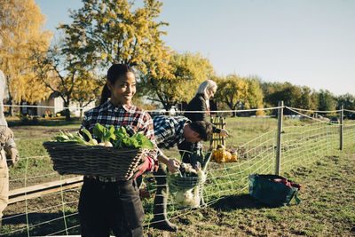 Smiling female farmer carrying basket of vegetables with friends in background at field