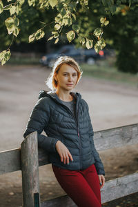 Portrait of woman wearing jacket leaning against fence while standing at park 