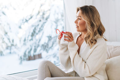 Young woman in white cardigan with cup of cocoa in hands looking at window with winter landscape 