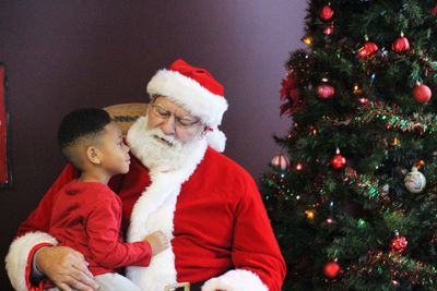 Santa claus sitting with cute boy on chair by christmas tree at home