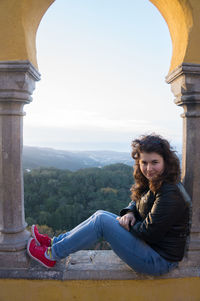 Side view portrait of woman sitting on retaining wall against sky