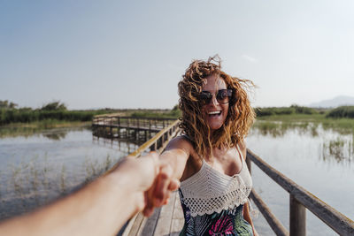 Woman holding hand of boyfriend while standing on footbridge against sky