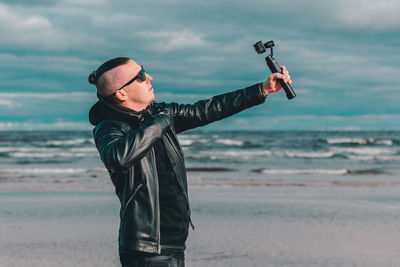 Young man wearing sunglasses filming with video camera against sea