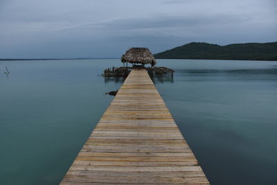 Jetty leading to thatched hut over river against cloudy sky