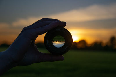 Human hand holding camera against sky during sunset