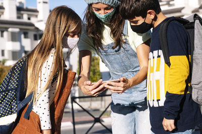 Mother and kids wearing protective face mask using smart phone standing in public park on sunny day