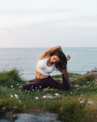 Full length of female athlete in sportswear sitting on grassy hill in eka pada raja kapotasana pose and looking down while practicing yoga in nature