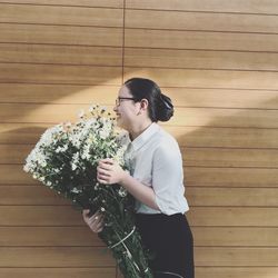 Young woman holding flower bouquet while standing against wooden wall