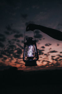 Cropped hand holding illuminated lantern against sky at sunset