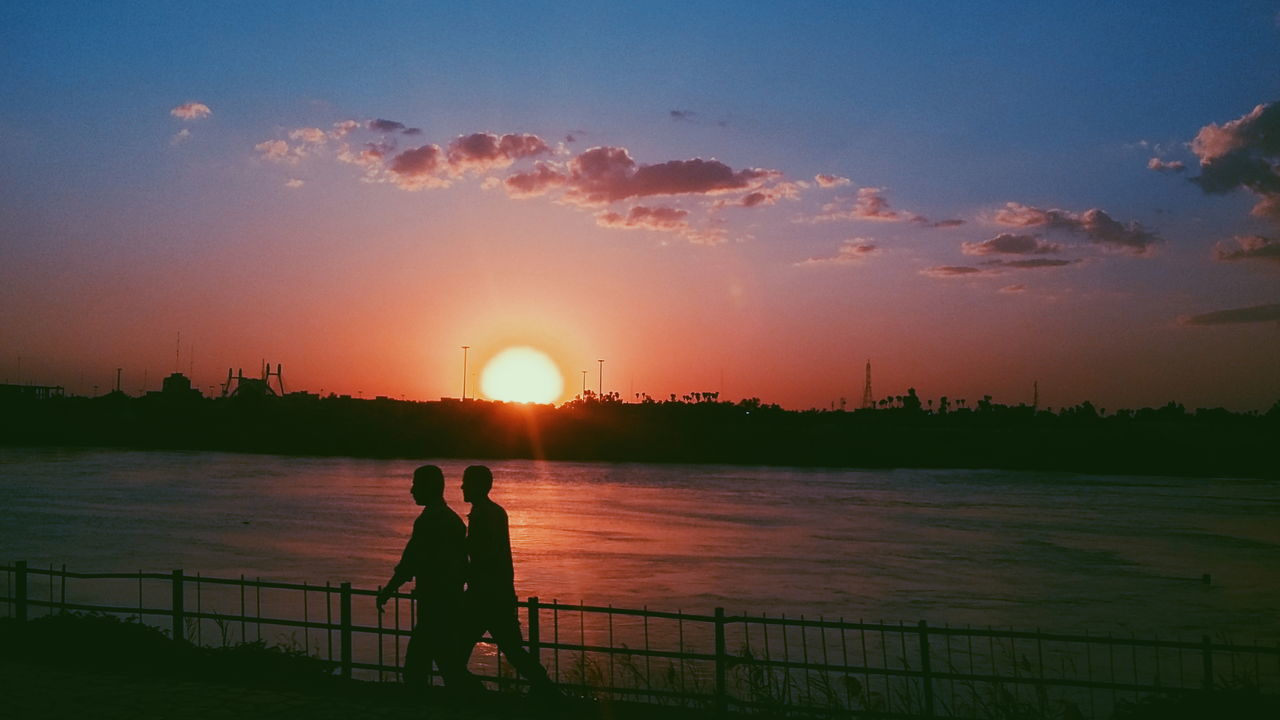 sunset, railing, sun, real people, water, two people, orange color, sky, nature, beauty in nature, sea, silhouette, men, love, sunlight, leisure activity, women, scenics, lifestyles, outdoors, togetherness, couple - relationship, built structure, standing, friendship, city, day, people