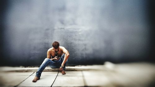 Full length of shirtless man using phone while sitting on footpath against wall