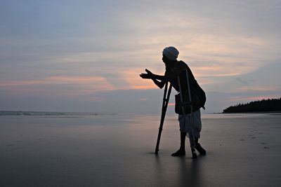 Full length of silhouette man with crutches standing at beach against sky during sunset