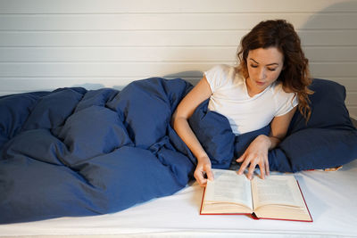 Young woman reading book while lying on bed at home