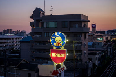 Road sign by illuminated buildings against sky at dusk