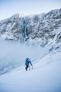 A male alpine climber ascends a steep section of snow with clouds and mountains behind