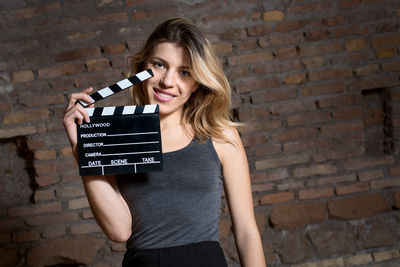 Portrait of young woman holding film slate while standing against brick wall