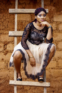 Portrait of young woman sitting on ladder against brick wall