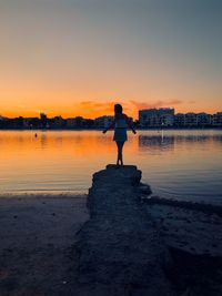 Rear view of woman standing at lakeshore against sky during sunset