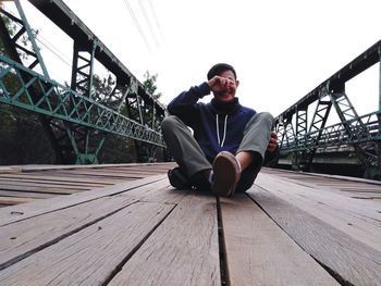 Young man sitting on footbridge against sky