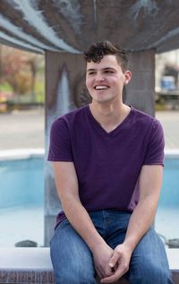 Smiling young man sitting on retaining wall against fountain
