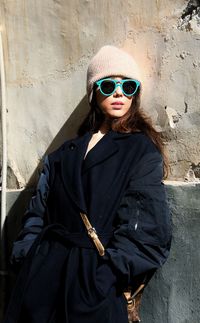 Portrait of young woman wearing sunglasses standing against wall