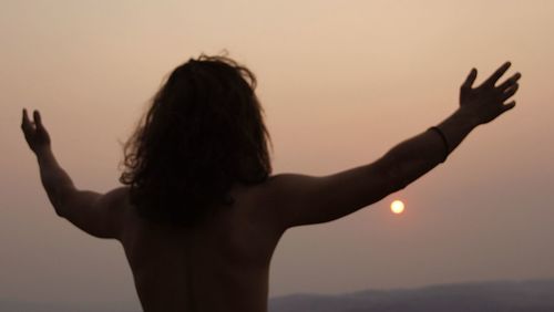 Rear view of shirtless woman standing against sky during sunset