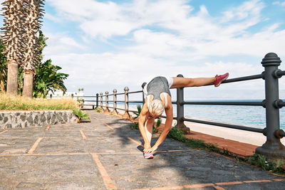 Young beautiful sportive woman training over seaside promenade, stretching legs before jogging. 