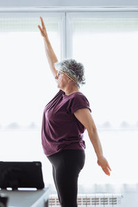 Side view of elderly female in activewear with short gray hair raising arm while doing stretching exercise against window in sunlit room in morning at home