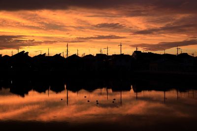 Silhouette houses by lake against orange sky