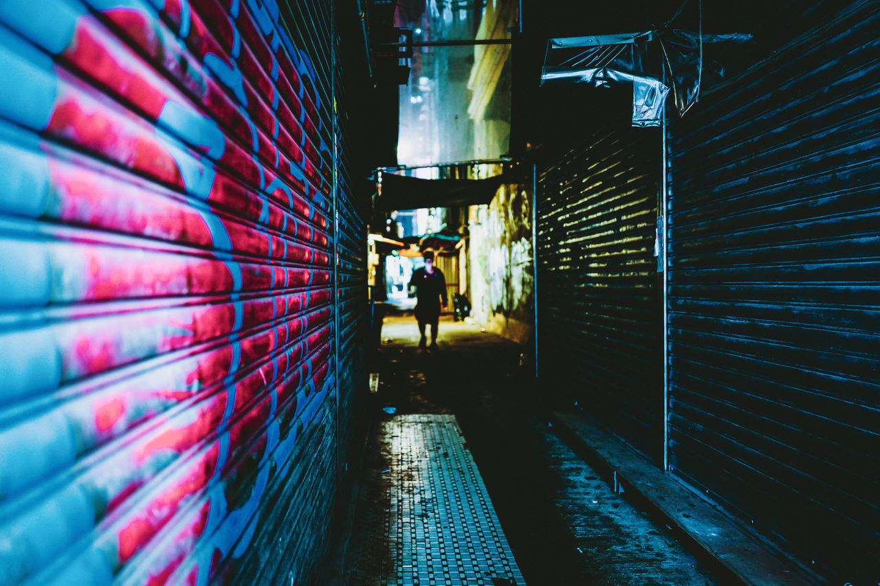 MAN WALKING ON FOOTPATH AGAINST ILLUMINATED BUILDING