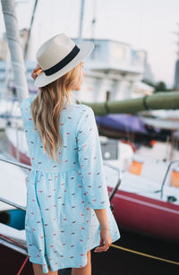 Beautiful blonde young woman in blue dress and straw hat on boat at pier in the sunset time