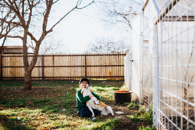 Young boy sitting outside backyard greehouse hugging corgi dog