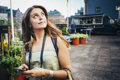 Thoughtful female tourist looking away while holding smart phone in city