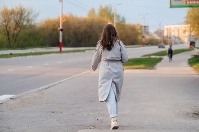 Girl with long hair in a grey trench coat outdoors on the street spring, view from the back